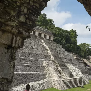 Stepped limestone pyramid with a temple on top, framed by a ruined stone arch, with a woman seated in the foreground.