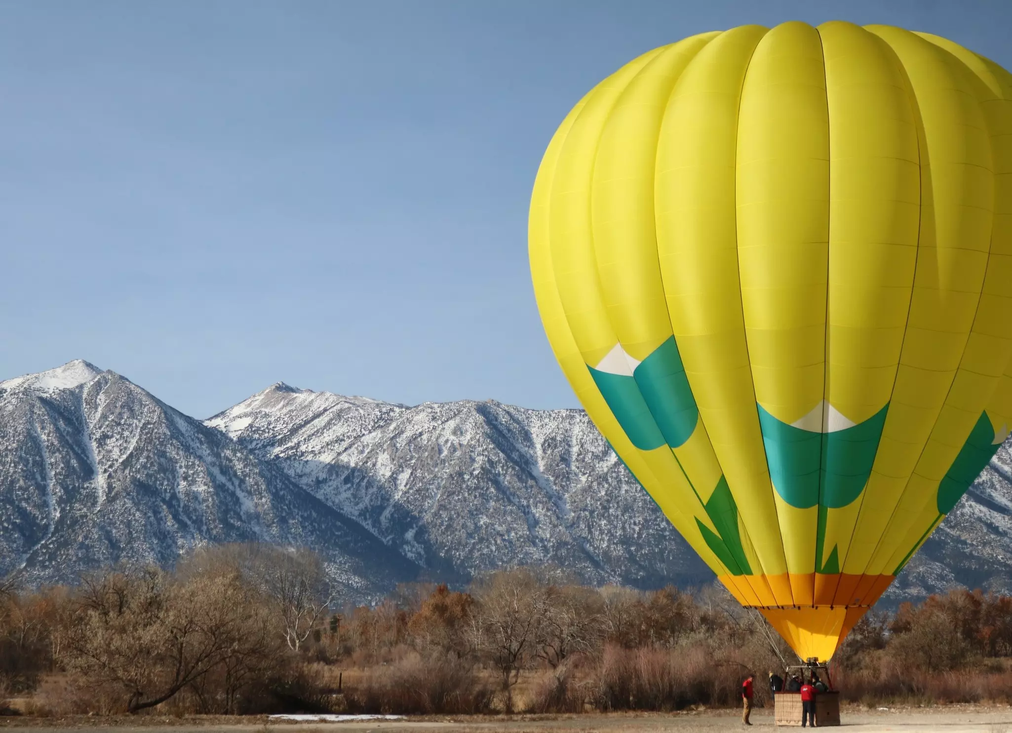 A yellow and green hot-air balloon prepares to take off from a plateau near some snow-capped mountains.