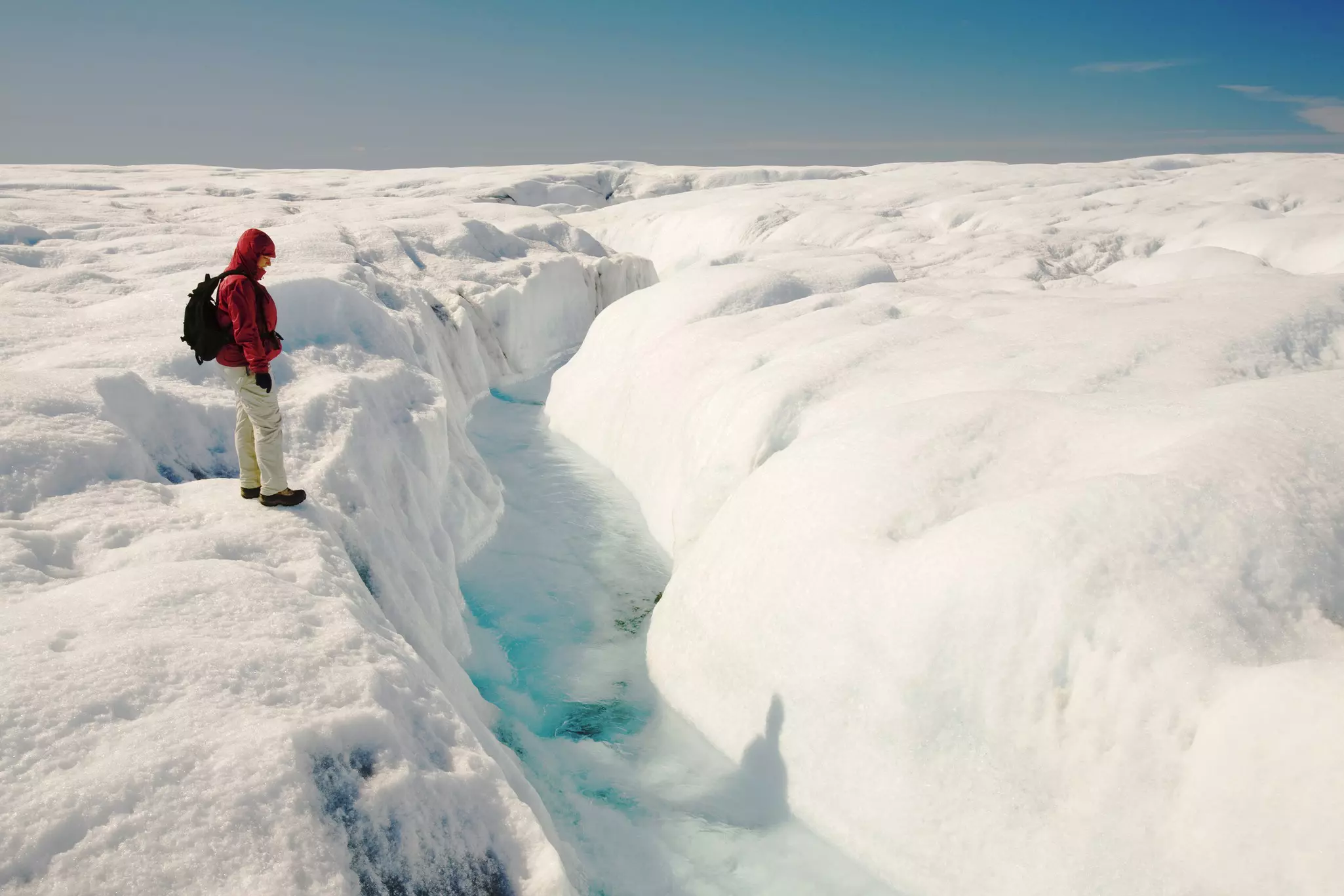 A person wearing a red jacket and black backpack stands on snow looking at a crevice of blue ice in Greenland.