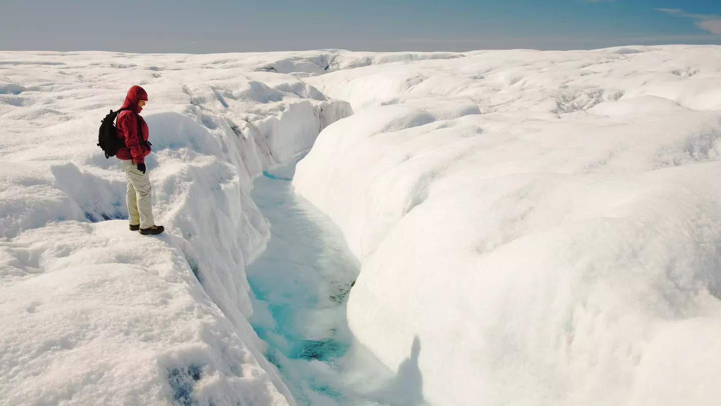 A person wearing a red jacket and black backpack stands on snow looking at a crevice of blue ice in Greenland.