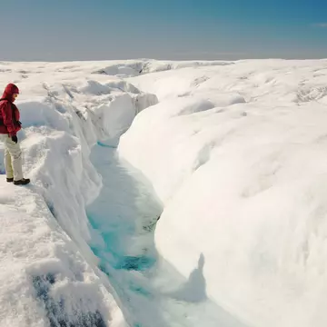 A person wearing a red jacket and black backpack stands on snow looking at a crevice of blue ice in Greenland.