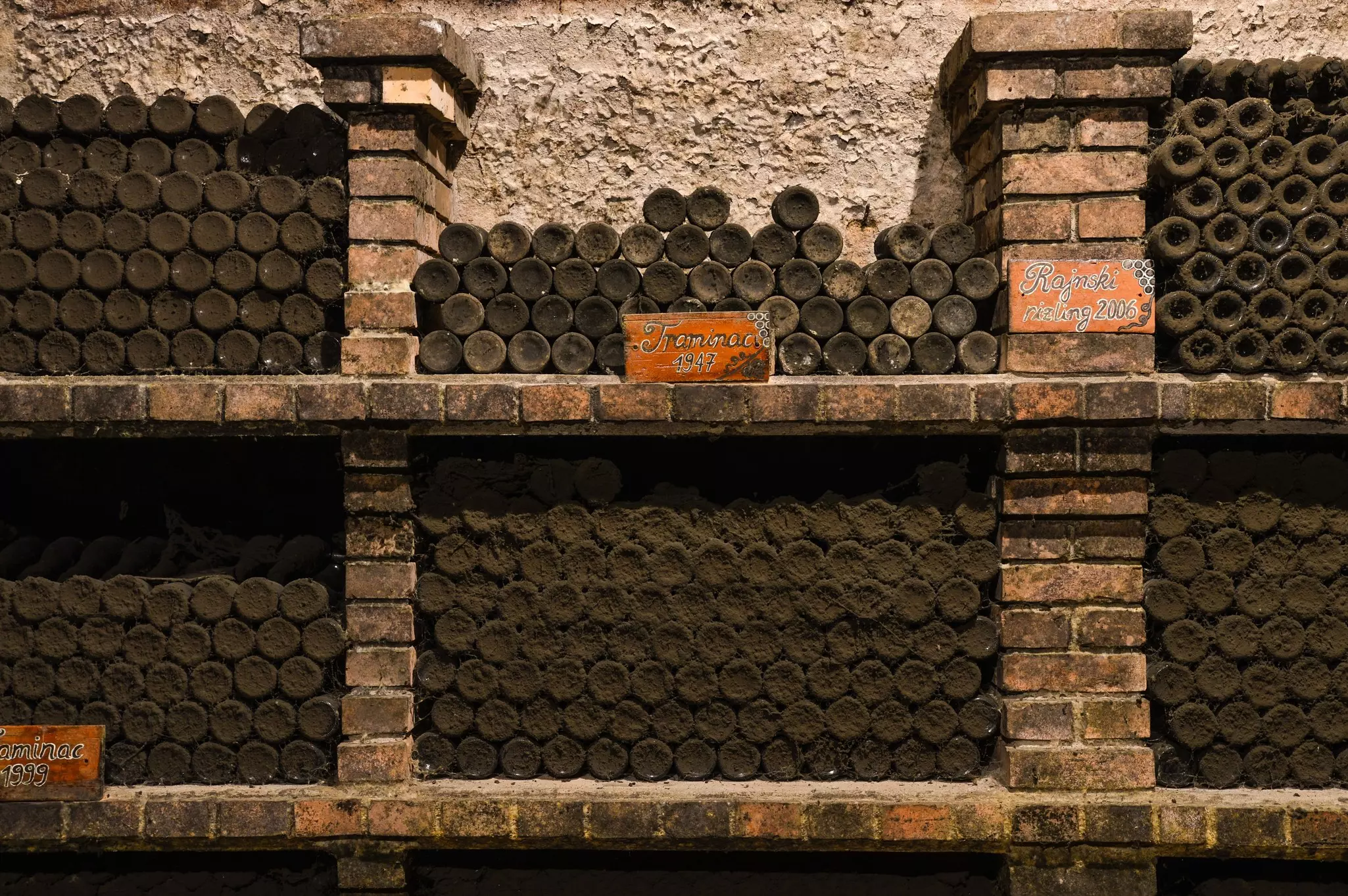 Dark, dusty wine bottles are stacked on their sides so only the bottoms are visible between brick shelves.