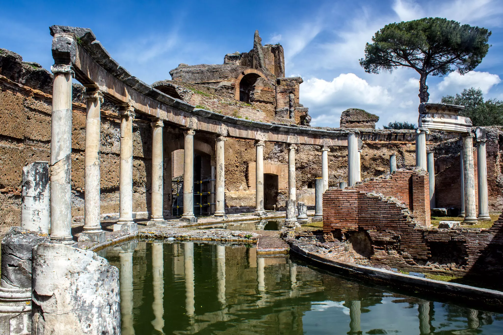 Exterior of the Roman ruins at Villa Adriana in Tivoli. A calm body of water fronts several pillars and the remains of a brick structure.