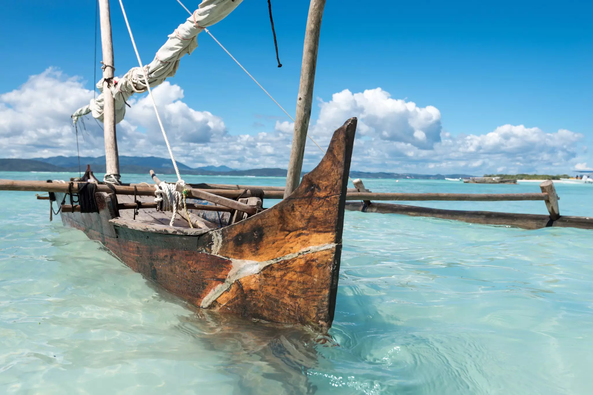 A wooden sailboat with pontoons is moored in crystal-clear water near a tropical beach.