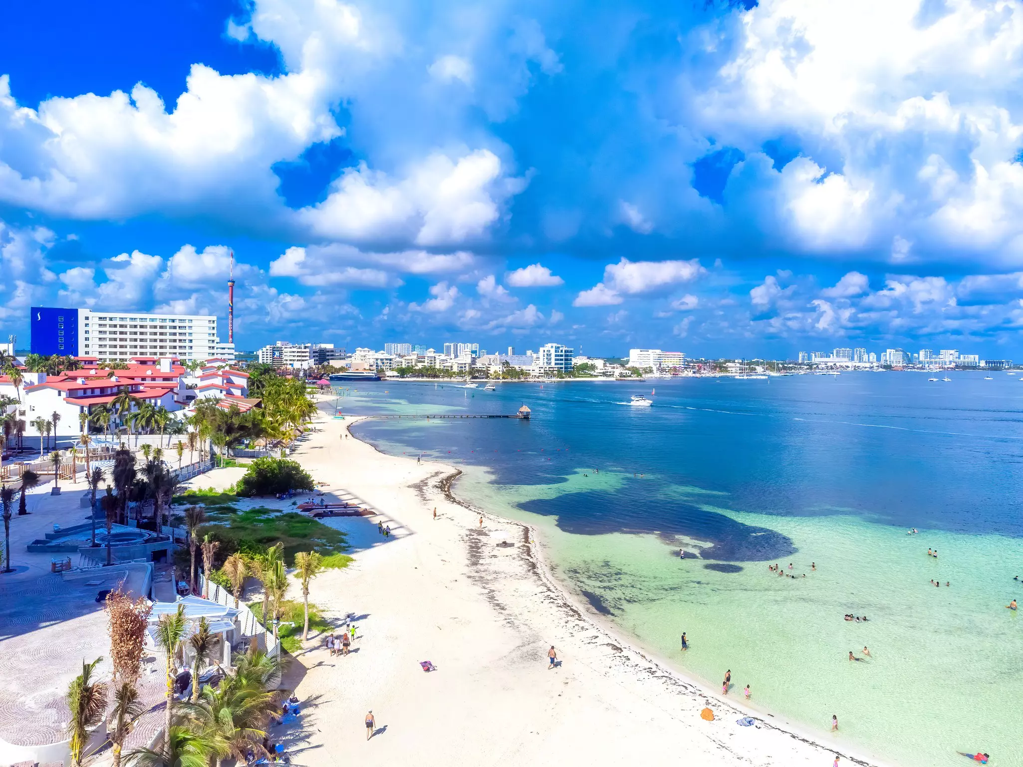 An aerial view of a beach with light-blue and -green waters. Palm trees and large hotels and resorts line the beach.