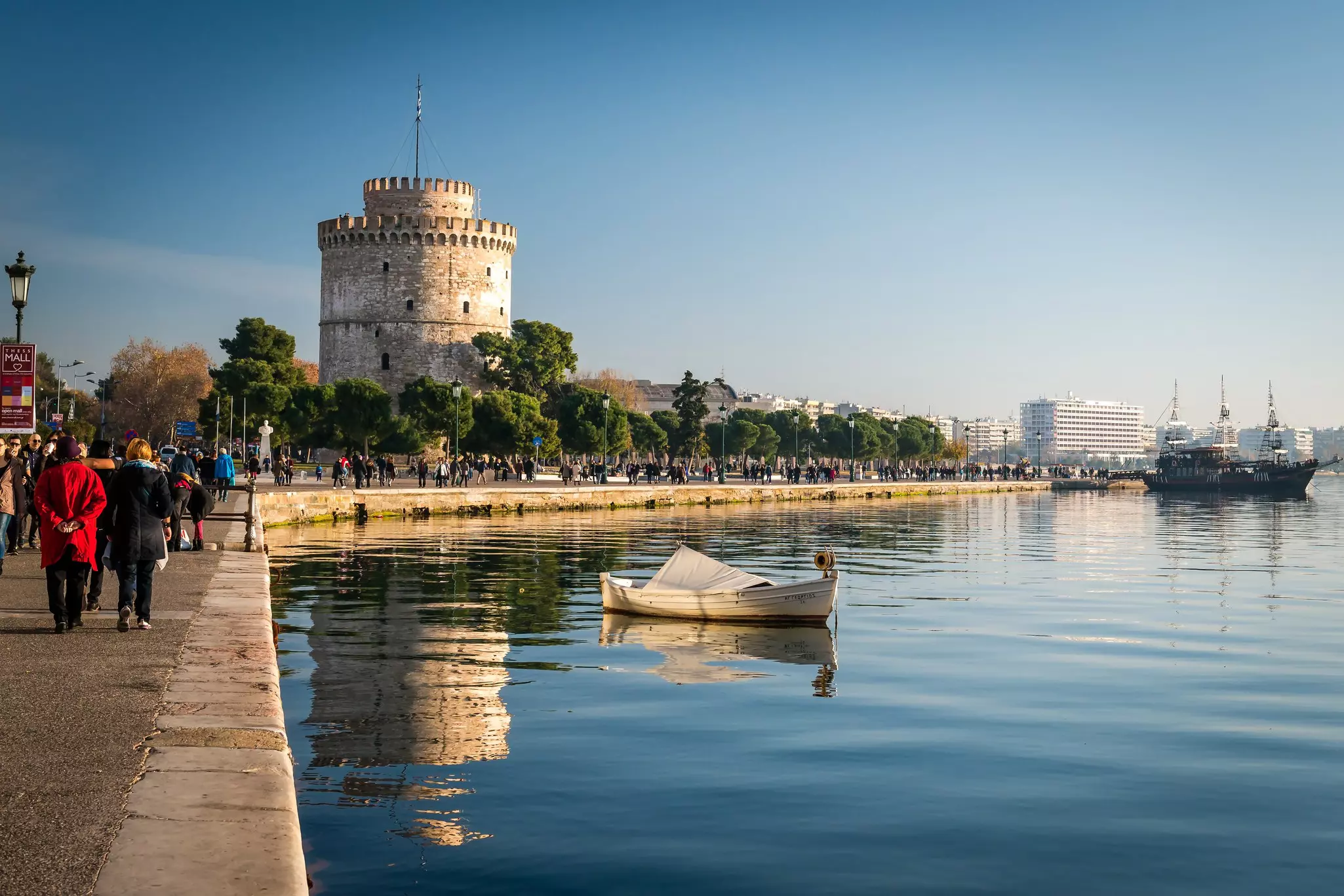 People walking along a waterfront near a wide, white tower