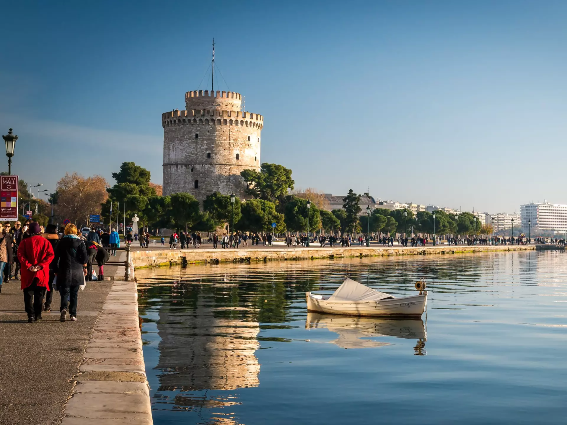 People walking along a waterfront near a wide, white tower
