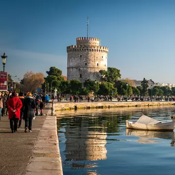 People walking along a waterfront near a wide, white tower