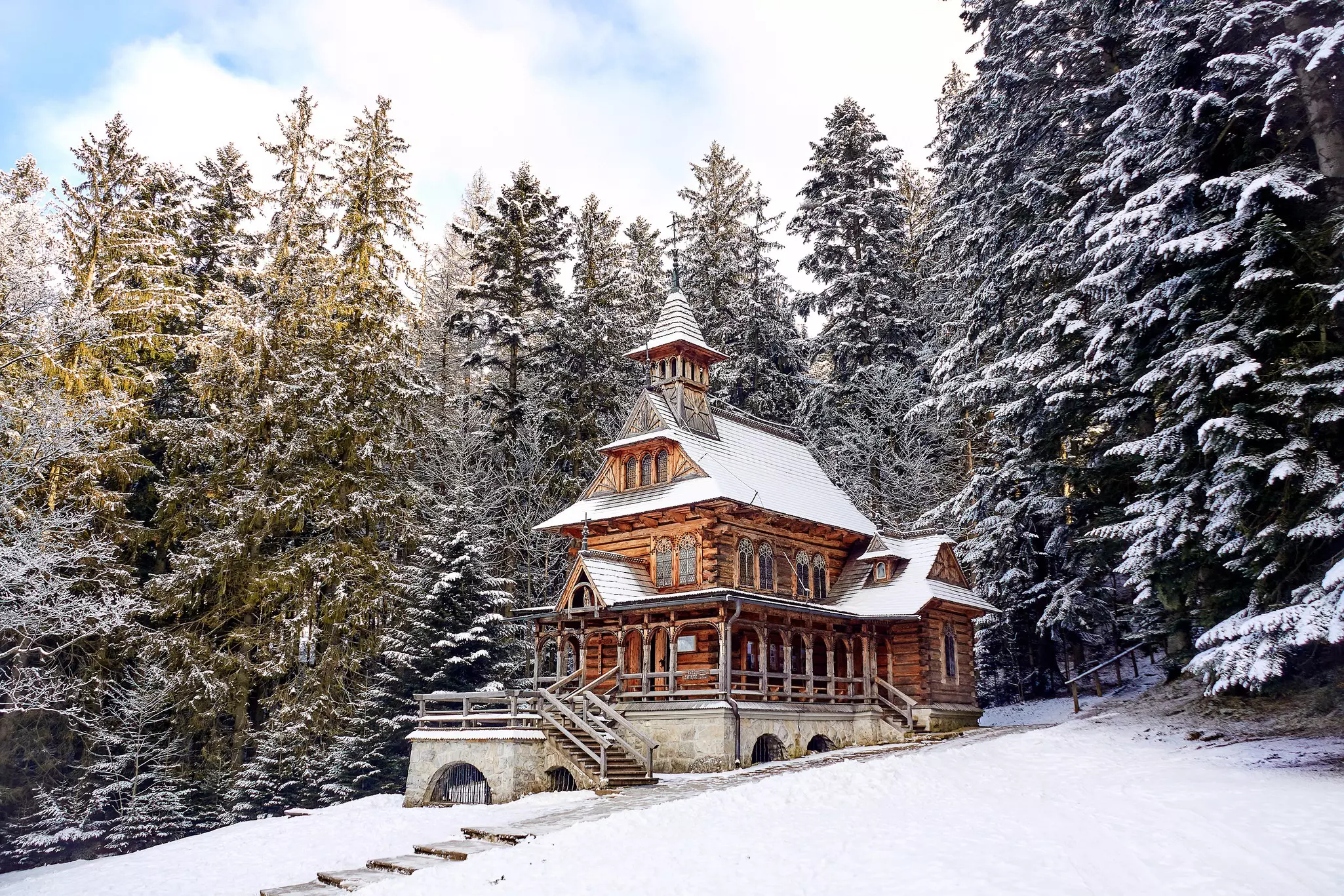 A wooden chapel with a steep peaked roof at the stop of a snowy incline, backed by evergreen trees dusted with snow.