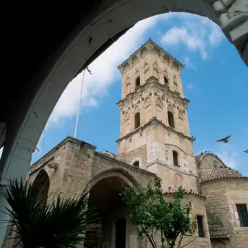 Carved stone bell tower rises above a domed church, framed by a shadowed stone arch against blue sky with birds in flight