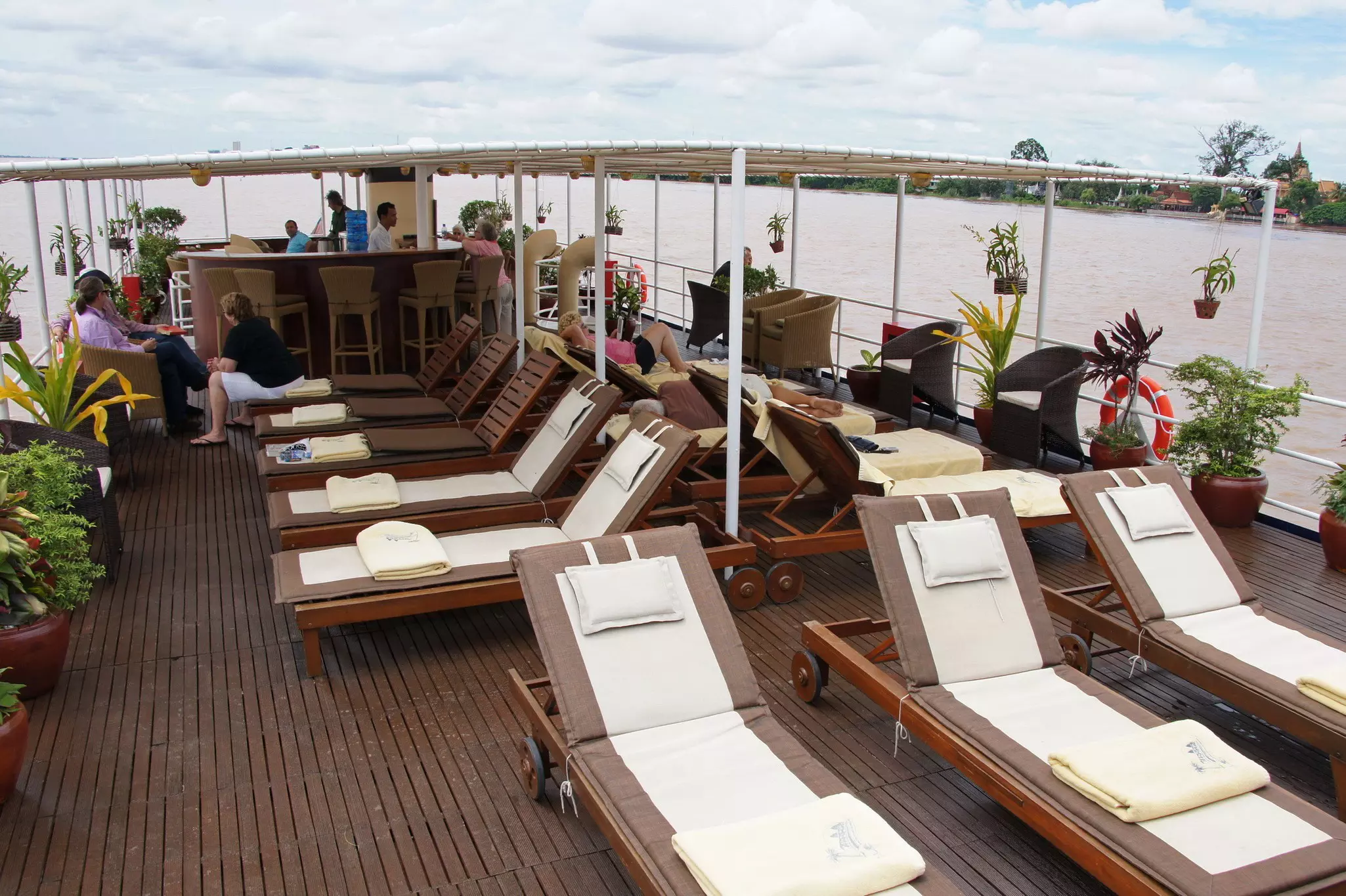 People relax in wicker chairs and on sunloungers on the covered wooden deck of a boat as it travels down a river.