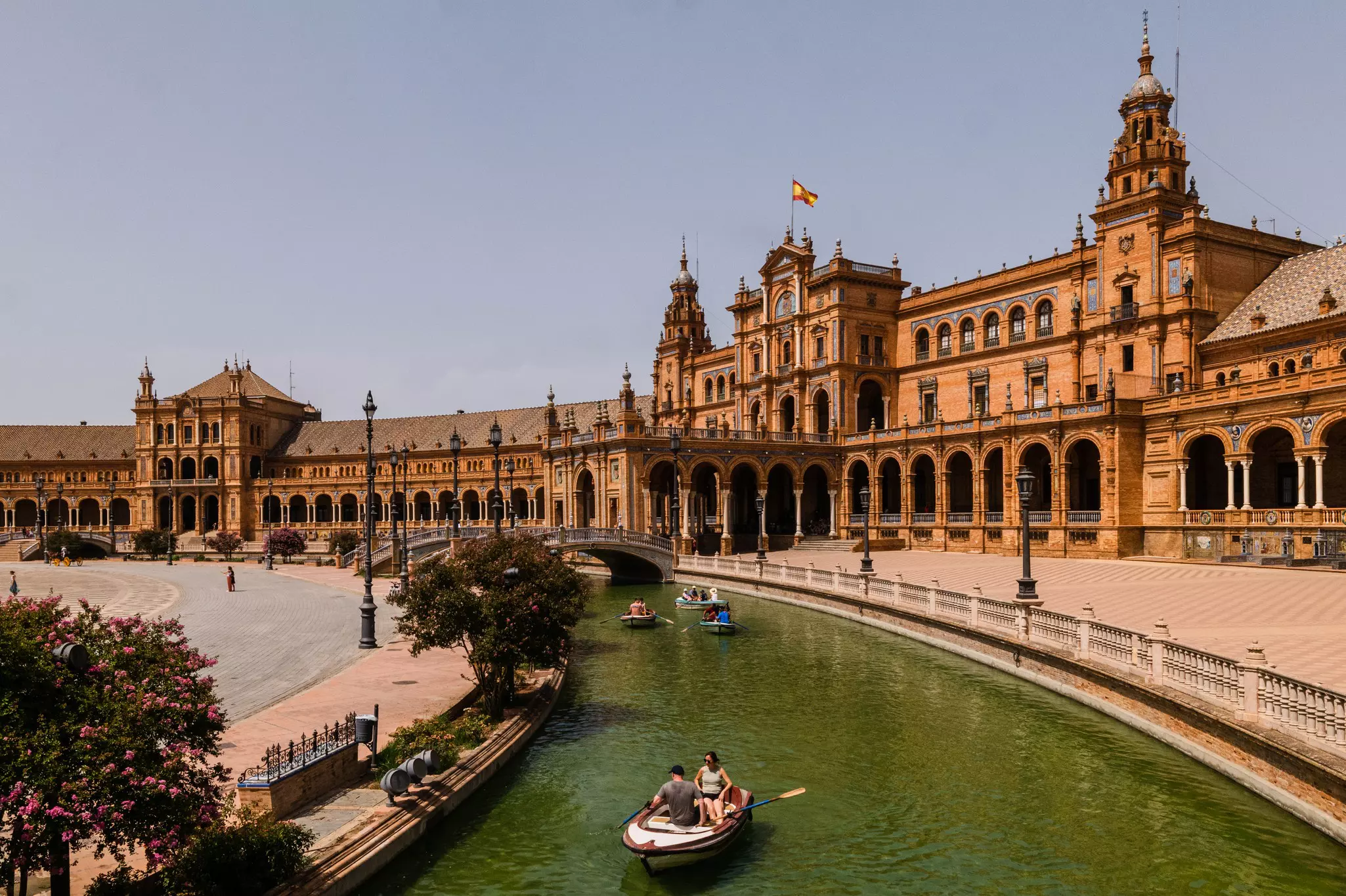 People in a boat on a waterway in a large plaza; there are arched bridges over the water. An elaborate red stone building fills one side of the plaza.