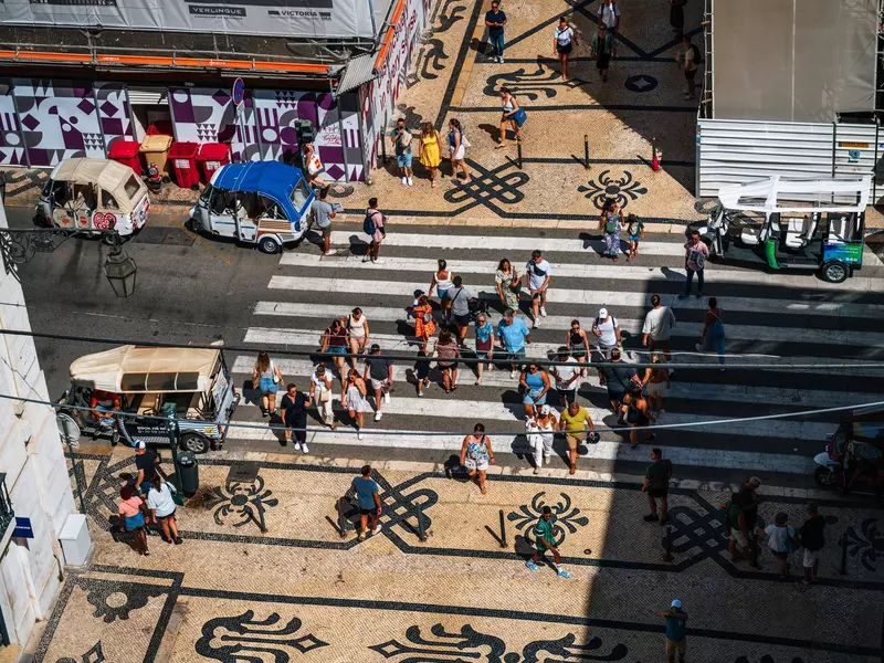 Bird's-eye view of pedestrians crossing a roadway through striped crosswalk. On either side of the street, an alley between city buildings has a decorative cobblestone path of white and black cobblestones.