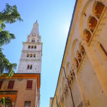 May 2022 Modena, Italy: Tower Ghirlandina and the facade of the Duomo Modena across the blue sky from beneath on the square piazza della Torre on a sunny day