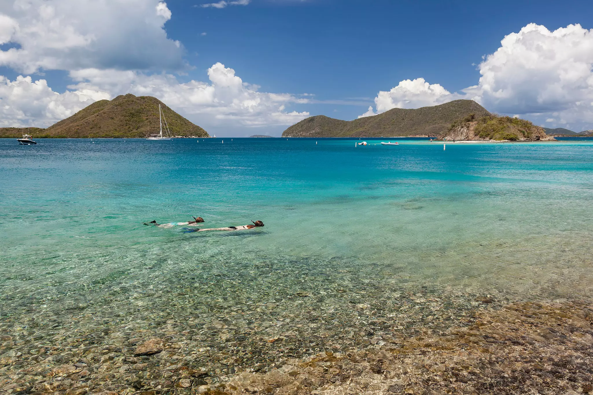 Two snorkelers explore clear waters just offshore with sail boats in the distance.