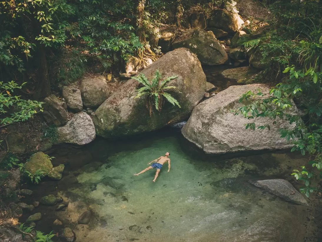 A man in swimwear lies in a natural pool gazing up at the rainforest that surrounds him