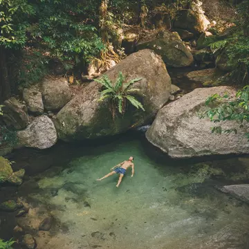 A man in swimwear lies in a natural pool gazing up at the rainforest that surrounds him