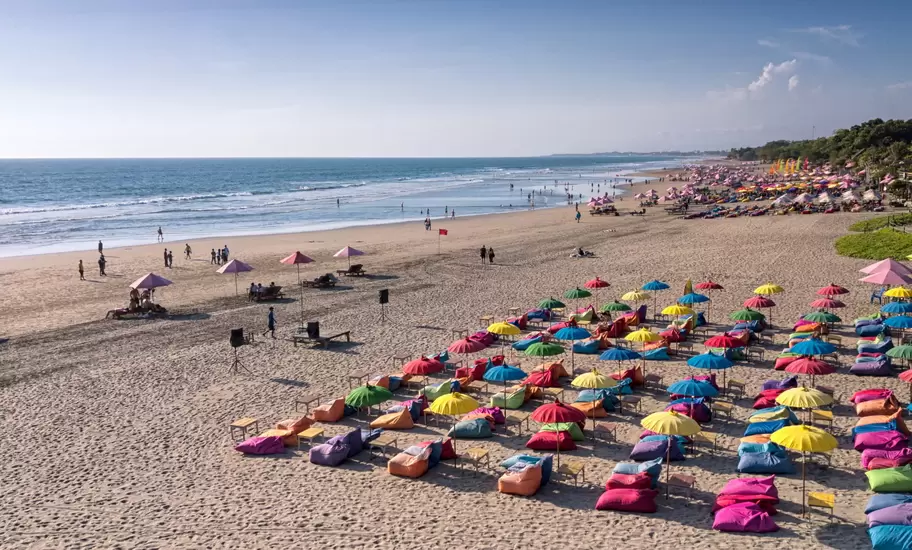 Wide sandy beach in Bali with rows of colourful umbrellas and beanbags, people strolling by the surf under a clear sky