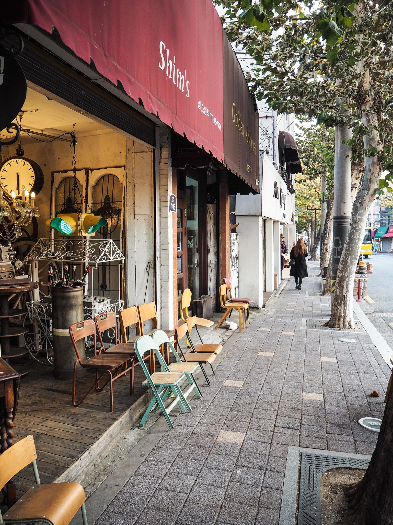 Mismatched vintage chairs outside an antique shop with a red awning reading Shim's, on a tree-lined pavement.