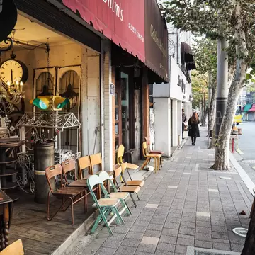 Mismatched vintage chairs outside an antique shop with a red awning reading Shim's, on a tree-lined pavement.