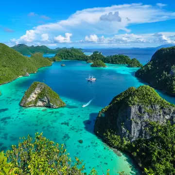 Jungle-covered karst islands ringing a turquoise lagoon at Wayag, Raja Ampat, with a sailing ship at anchor