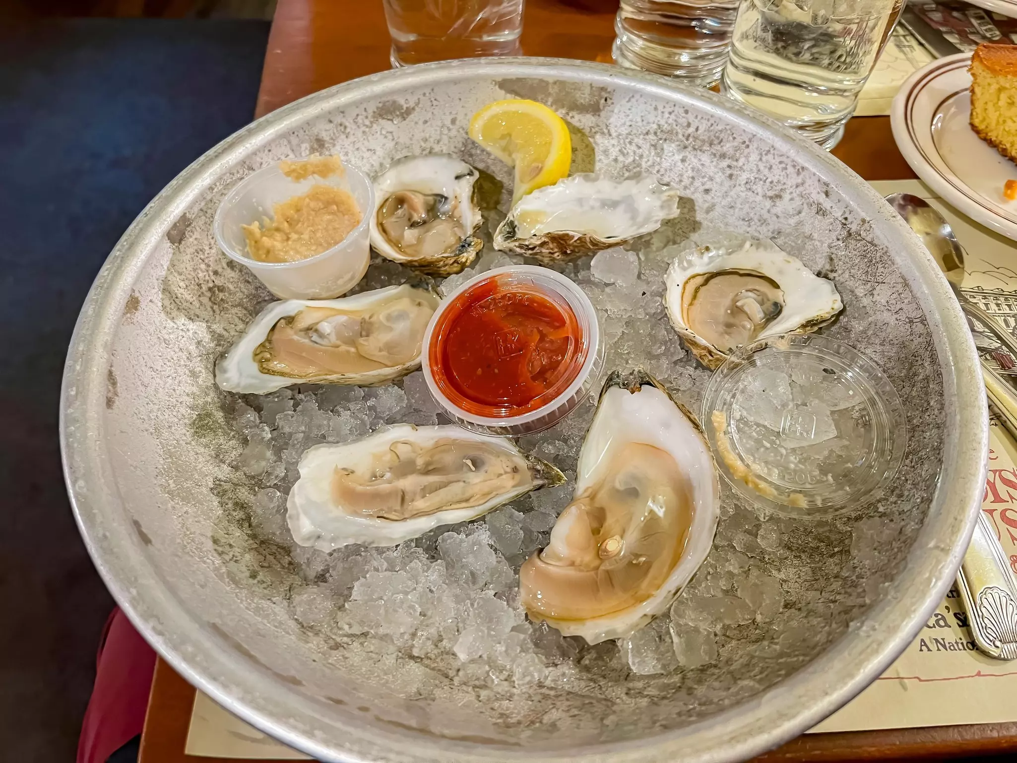 A large dish with six oysters served on ice with a piece of lemon and two small pots of sauce.