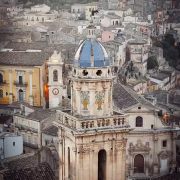 A bell tower with a blue-tiled cupola and ceramic panels in Ragusa Ibla, Sicily, above stone houses climbing a hill at dusk.