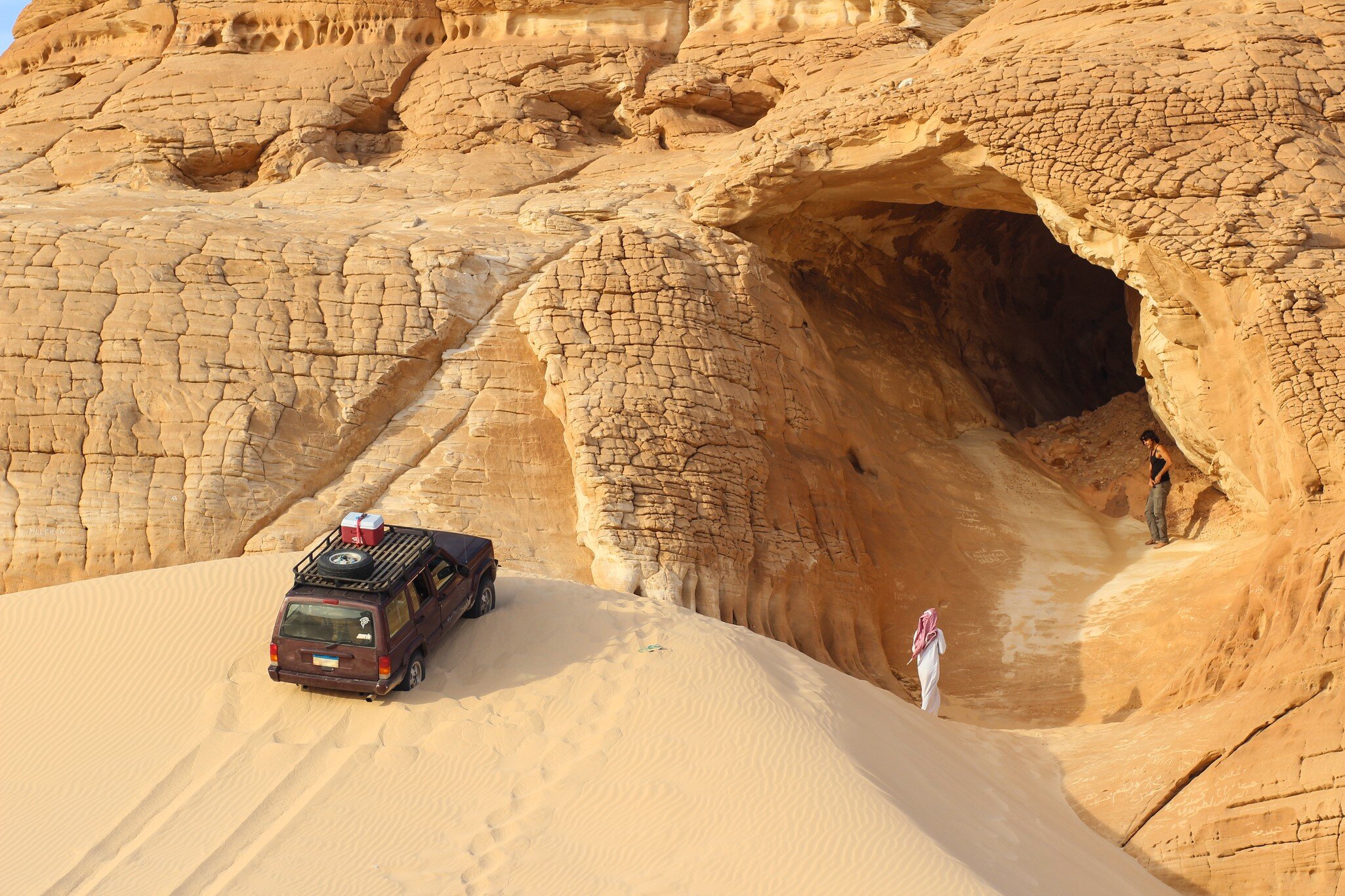 Spectacular Sinai Desert landscape (Gilf Kebir Plateau in Egypt). Thin sand dune leading to a cage in yellow sandstone mountain. Car on the dune top, a man in the cage, a bedouin man walking.; Shutterstock ID 690954682;
