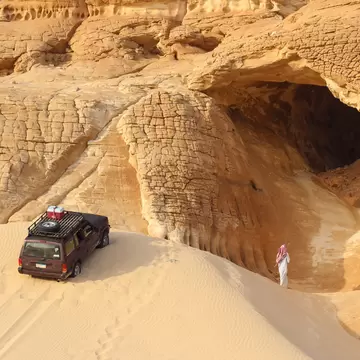Spectacular Sinai Desert landscape (Gilf Kebir Plateau in Egypt). Thin sand dune leading to a cage in yellow sandstone mountain. Car on the dune top, a man in the cage, a bedouin man walking.; Shutterstock ID 690954682;