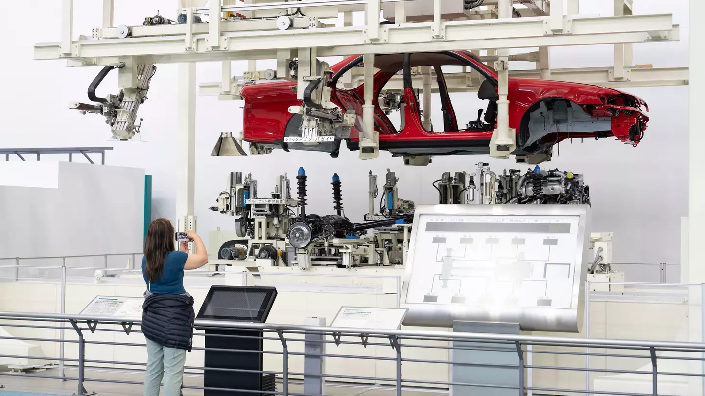 A woman takes a photo of an assembly line with a red car chassis held above the internal parts of the car.