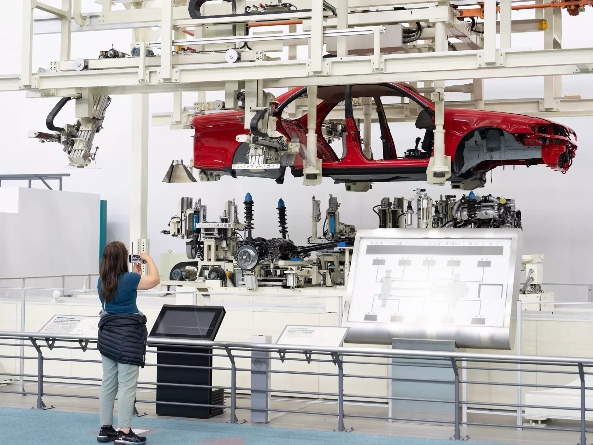 A woman takes a photo of an assembly line with a red car chassis held above the internal parts of the car.