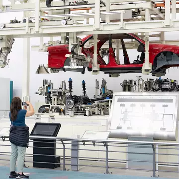 A woman takes a photo of an assembly line with a red car chassis held above the internal parts of the car.