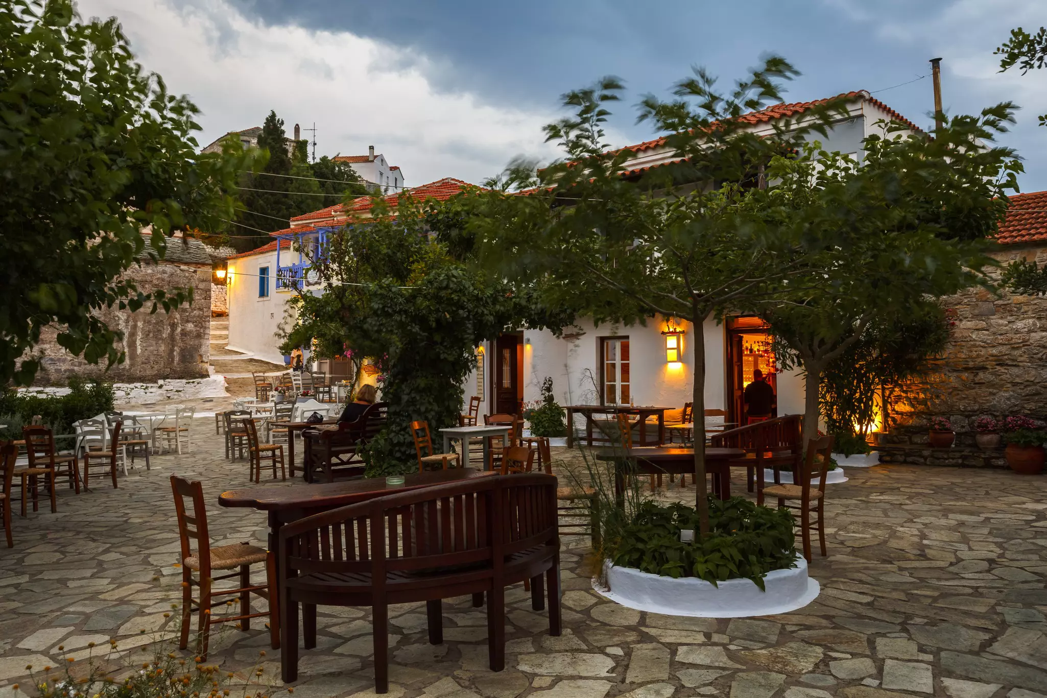 Empty tables in a cobblestone square in front of a white building with trees intertwined in front of the entrance on an island in the northern Sporades, Greece.