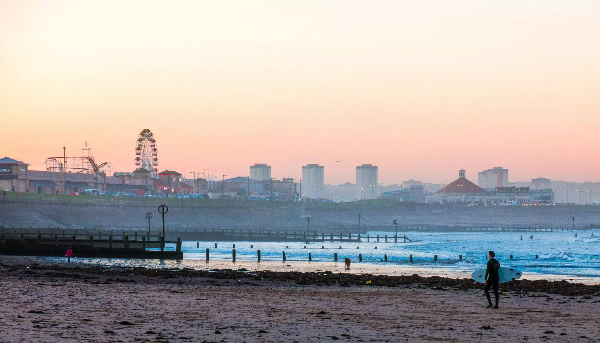 A man in silhouette carries a surfboard on a beach with a city skyline including a large Ferris wheel