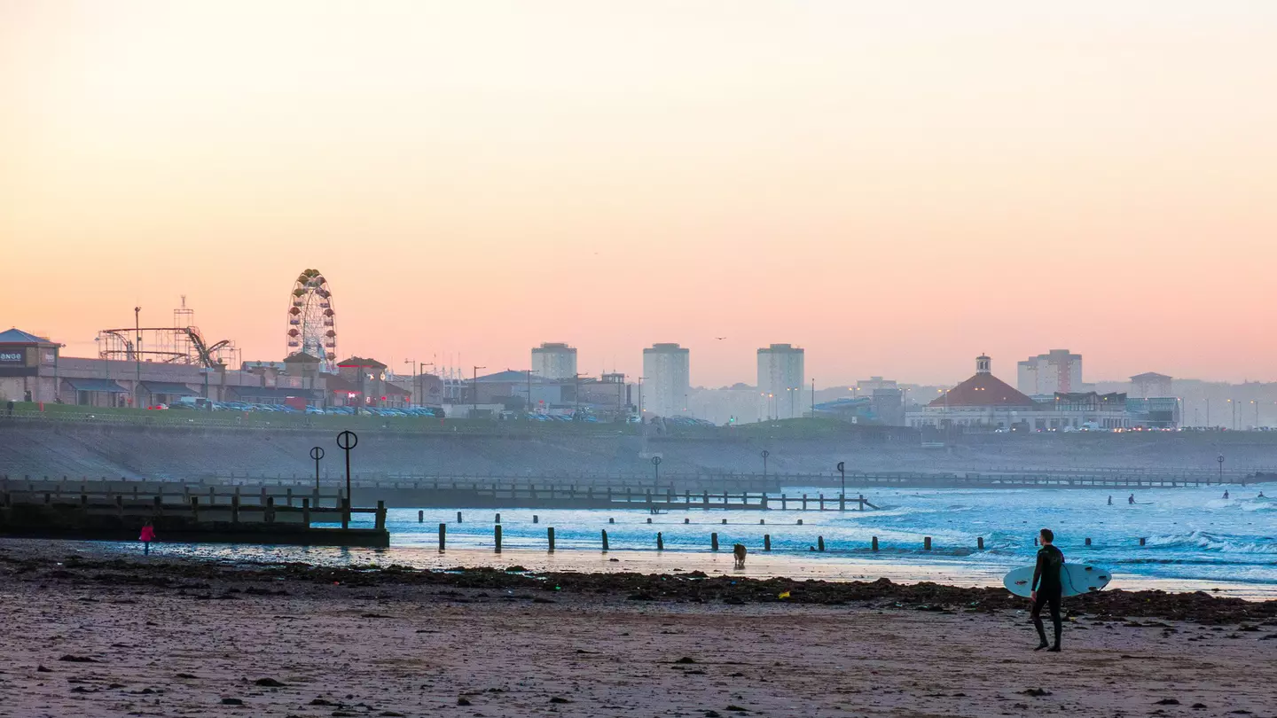 A man in silhouette carries a surfboard on a beach with a city skyline including a large Ferris wheel