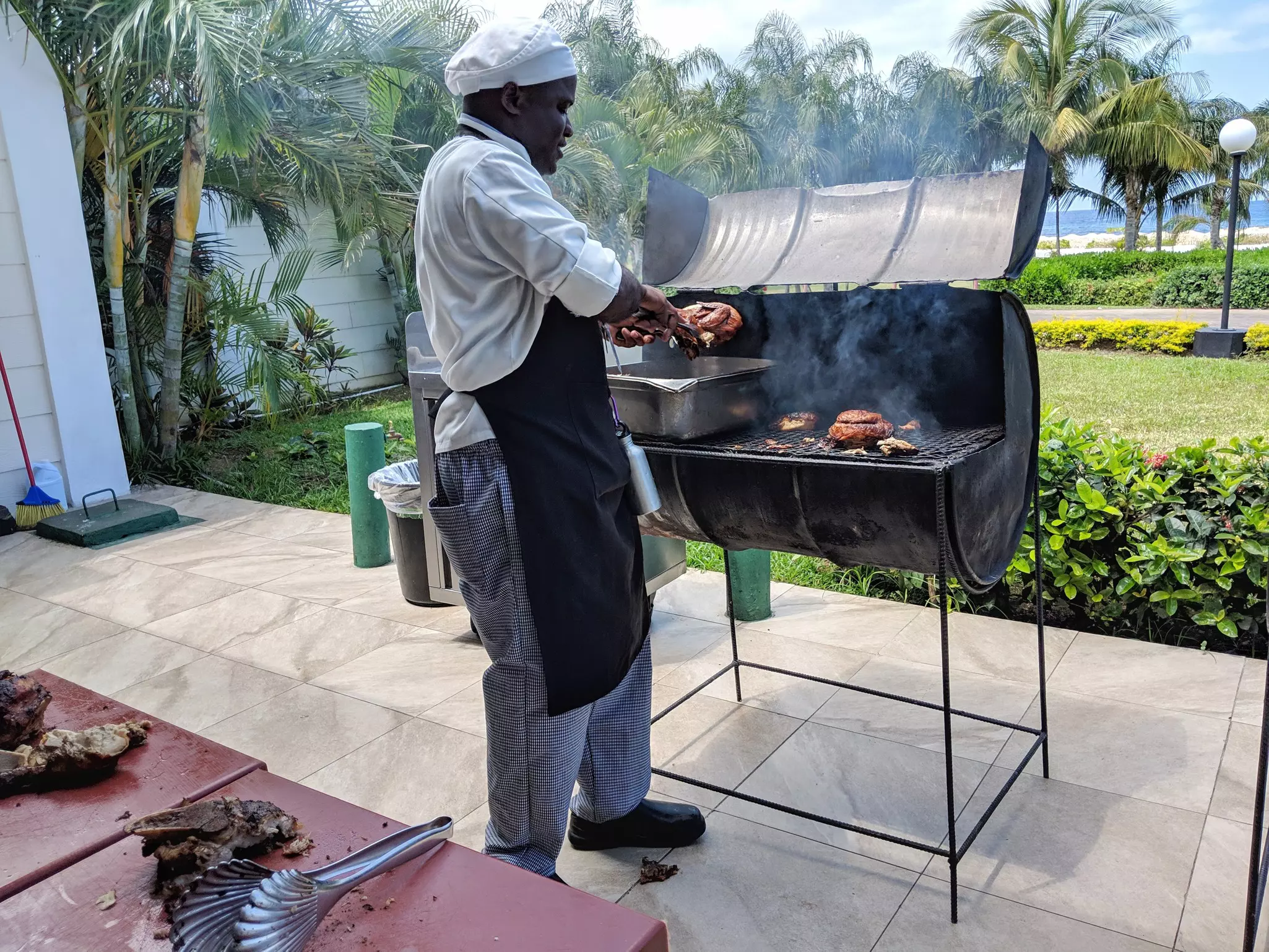 A chef cooks jerk chicken on a traditional oil drum grill in front of palm trees, a lawn and the sea.