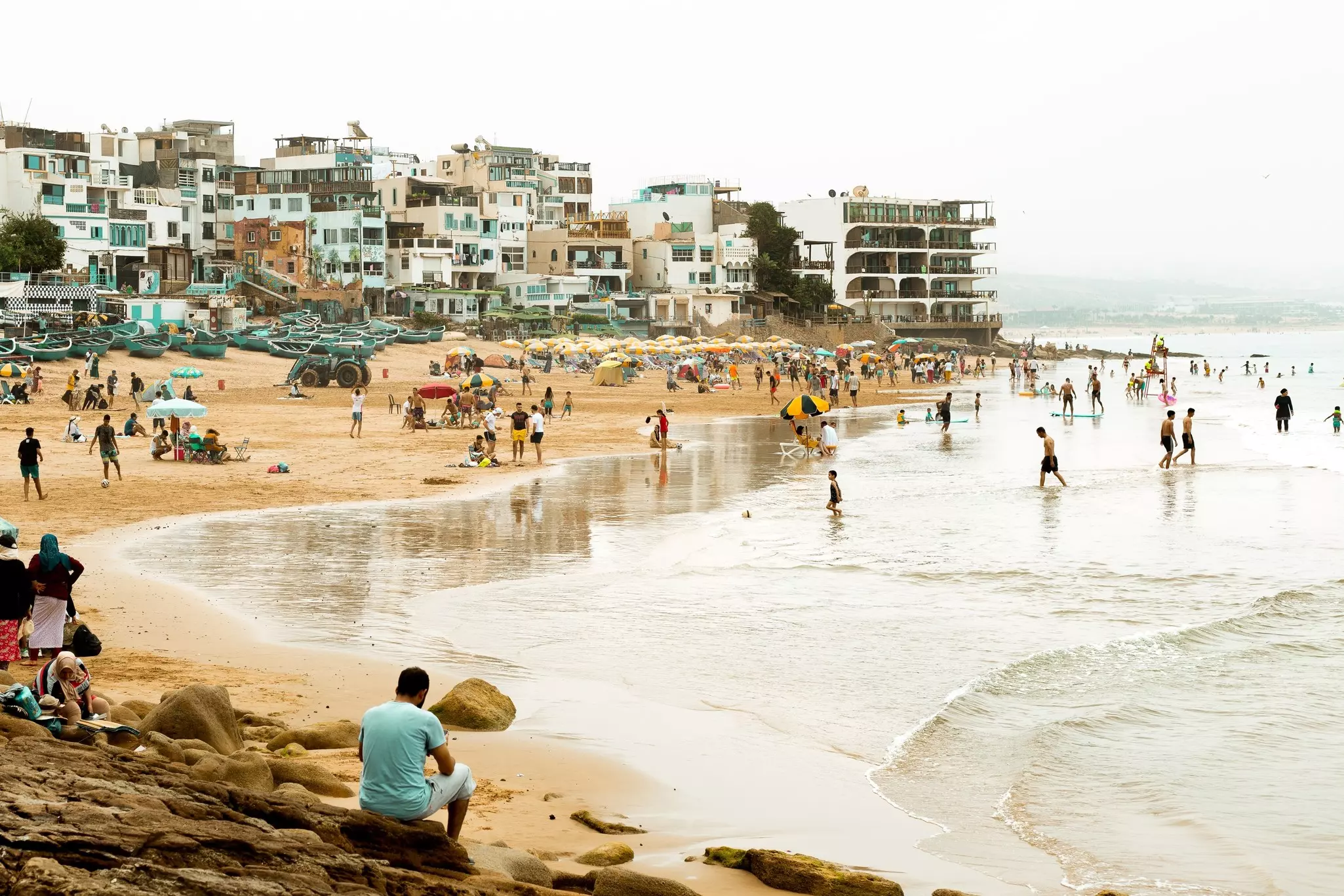 People on a golden sand beach, in the water and sitting on rocks in the foreground, with many multistory buildings bordering the beach.