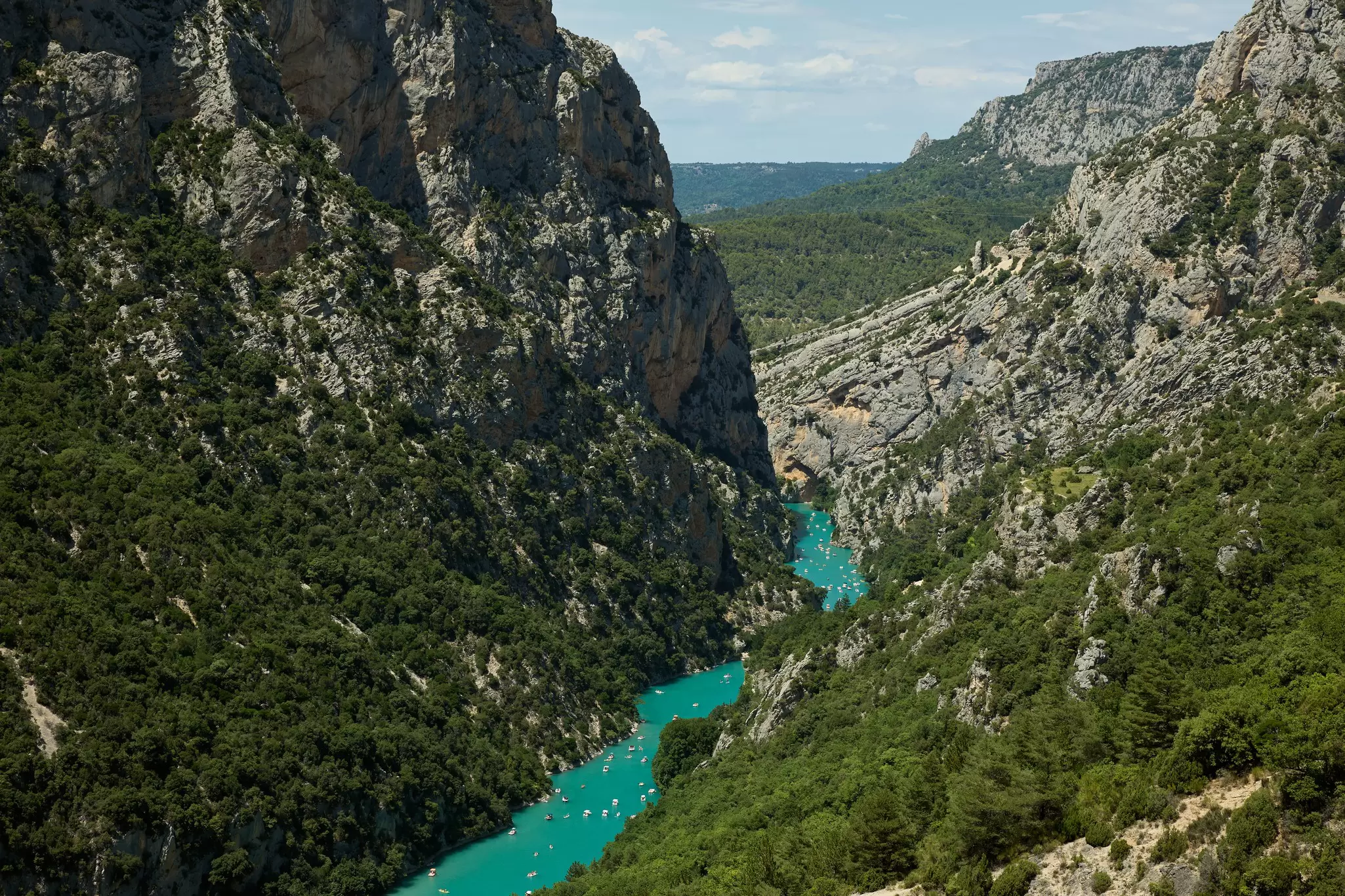 A turquoise river through a steep tree-lined gorge has small boats floating in it.