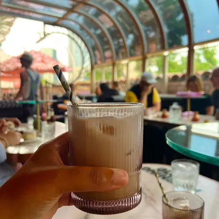 A hand holding a glass cup of iced coffee with a straw with out-of-focus patrons at tables in the background.