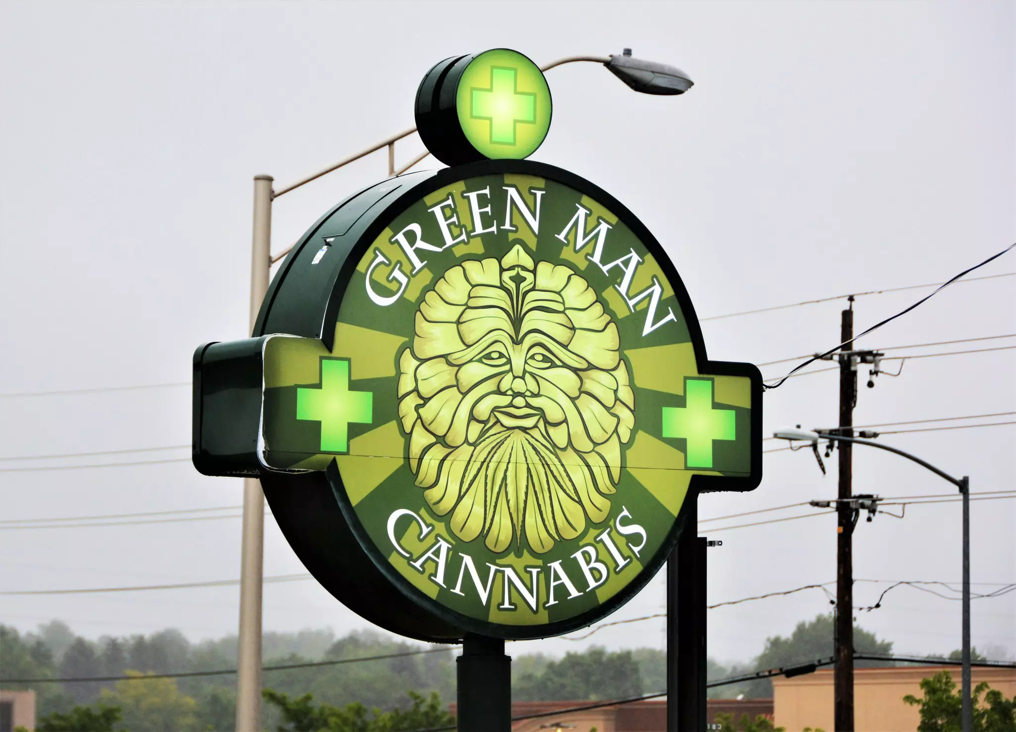 A middle aged white woman with short blond hair in a blue floral tunic, black slacks, and black sandals speaks with two budtenders at a Denver, Colorado cannabis dispensary. A long display case and counter made of blonde wood and glass shows cases a variety of edible and topical cannabis products and concentrates, while jars of flower sit on shelves on the wall