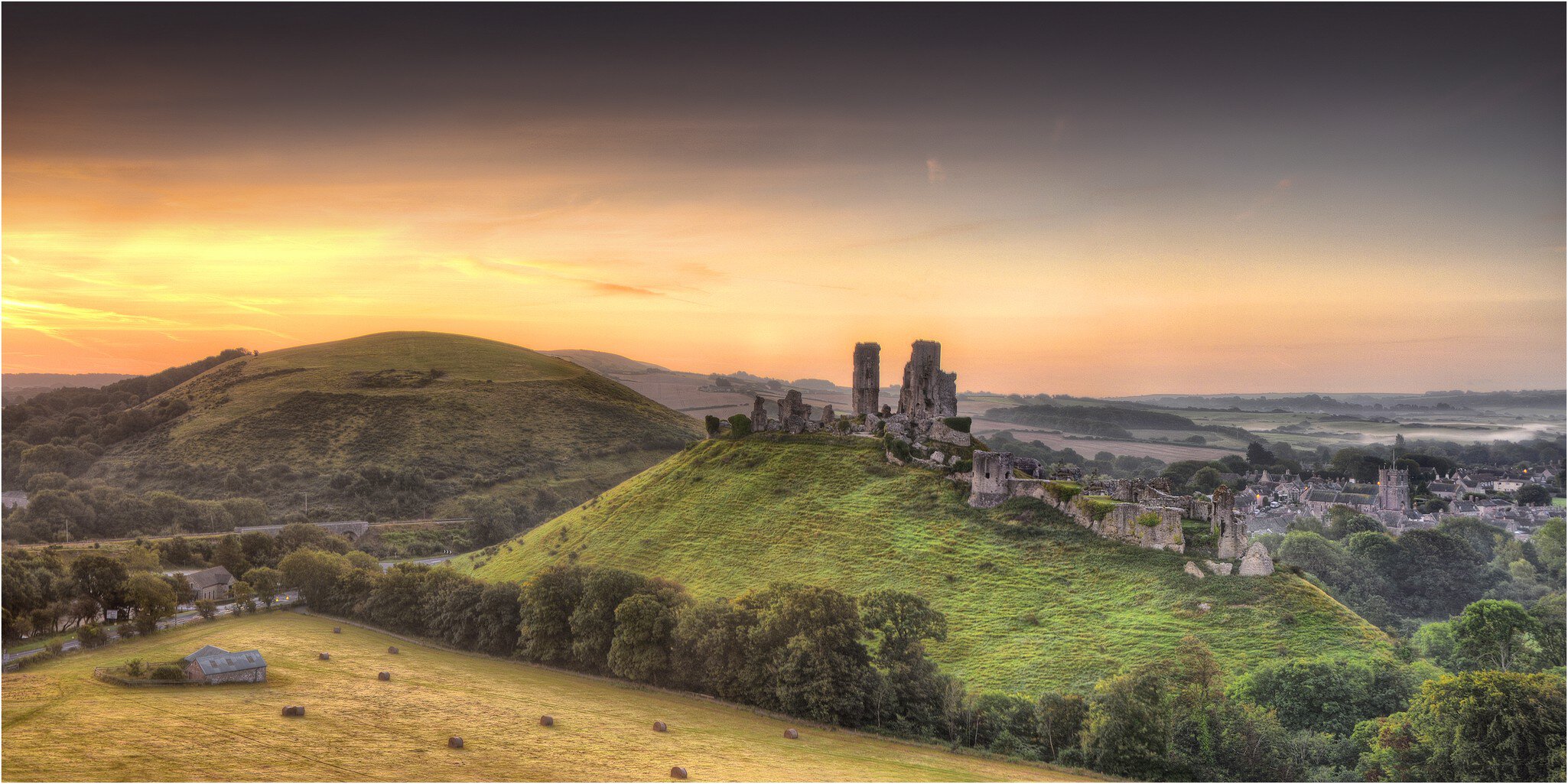 Ruined stone castle towers on a green hilltop above a village at golden sunset, with hay bales in the fields below