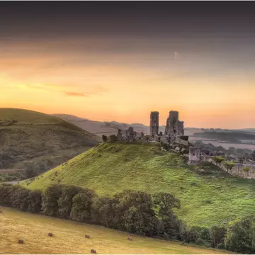 Ruined stone castle towers on a green hilltop above a village at golden sunset, with hay bales in the fields below
