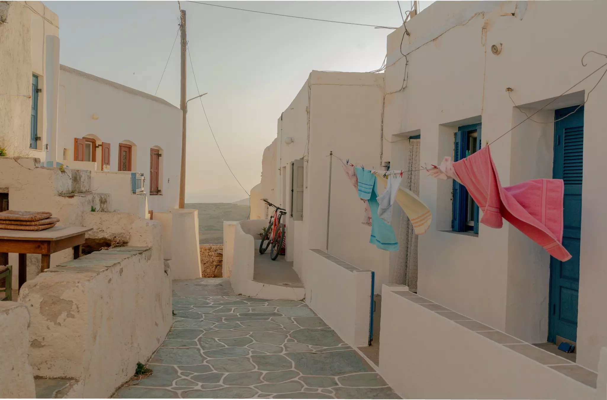 Colorful towels hang on the line outside a white building on a narrow cobblestone walkway in the Cyclades, Greece; the sea is visible at the end of the alley.