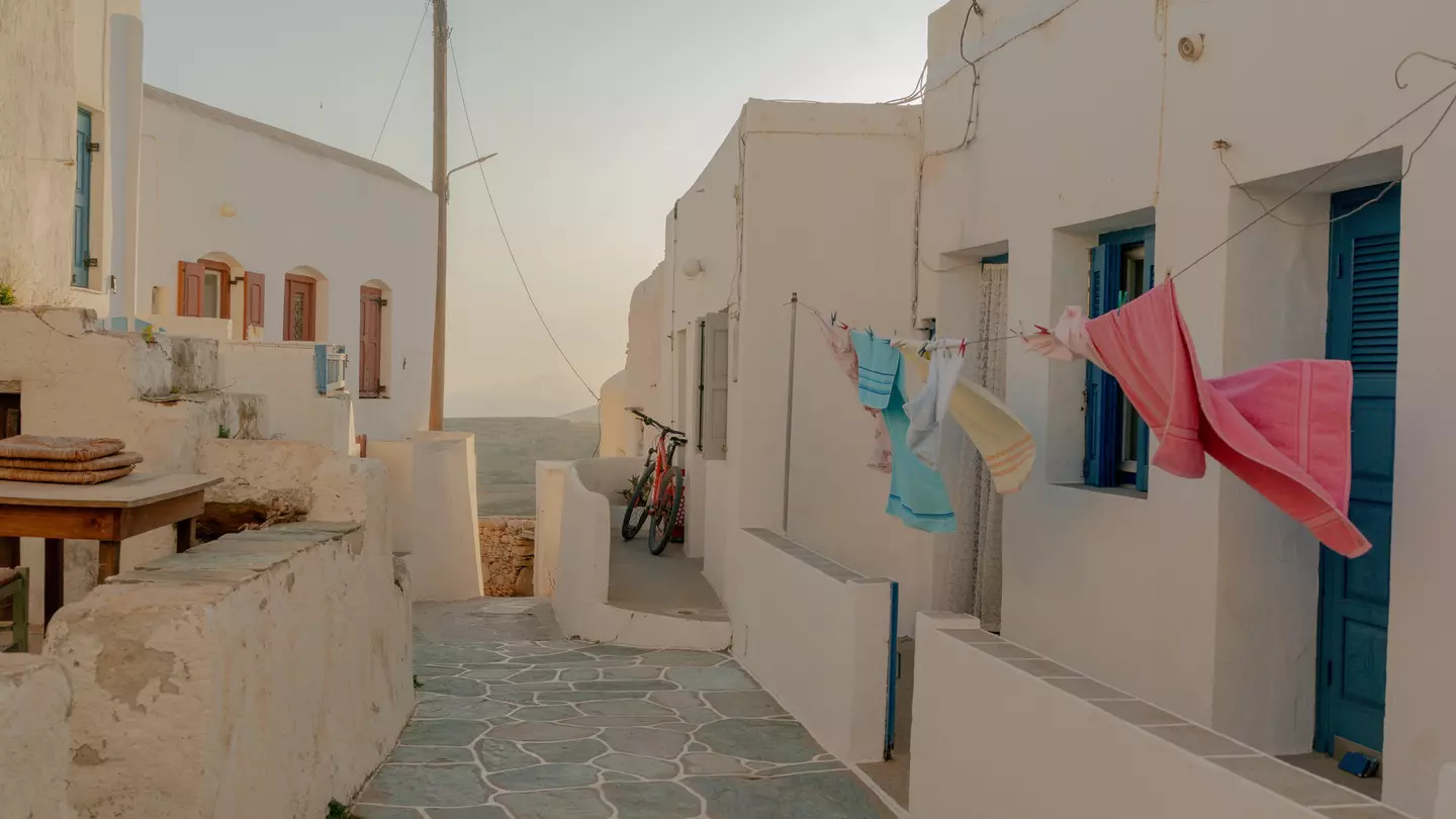 Colorful towels hang on the line outside a white building on a narrow cobblestone walkway in the Cyclades, Greece; the sea is visible at the end of the alley.