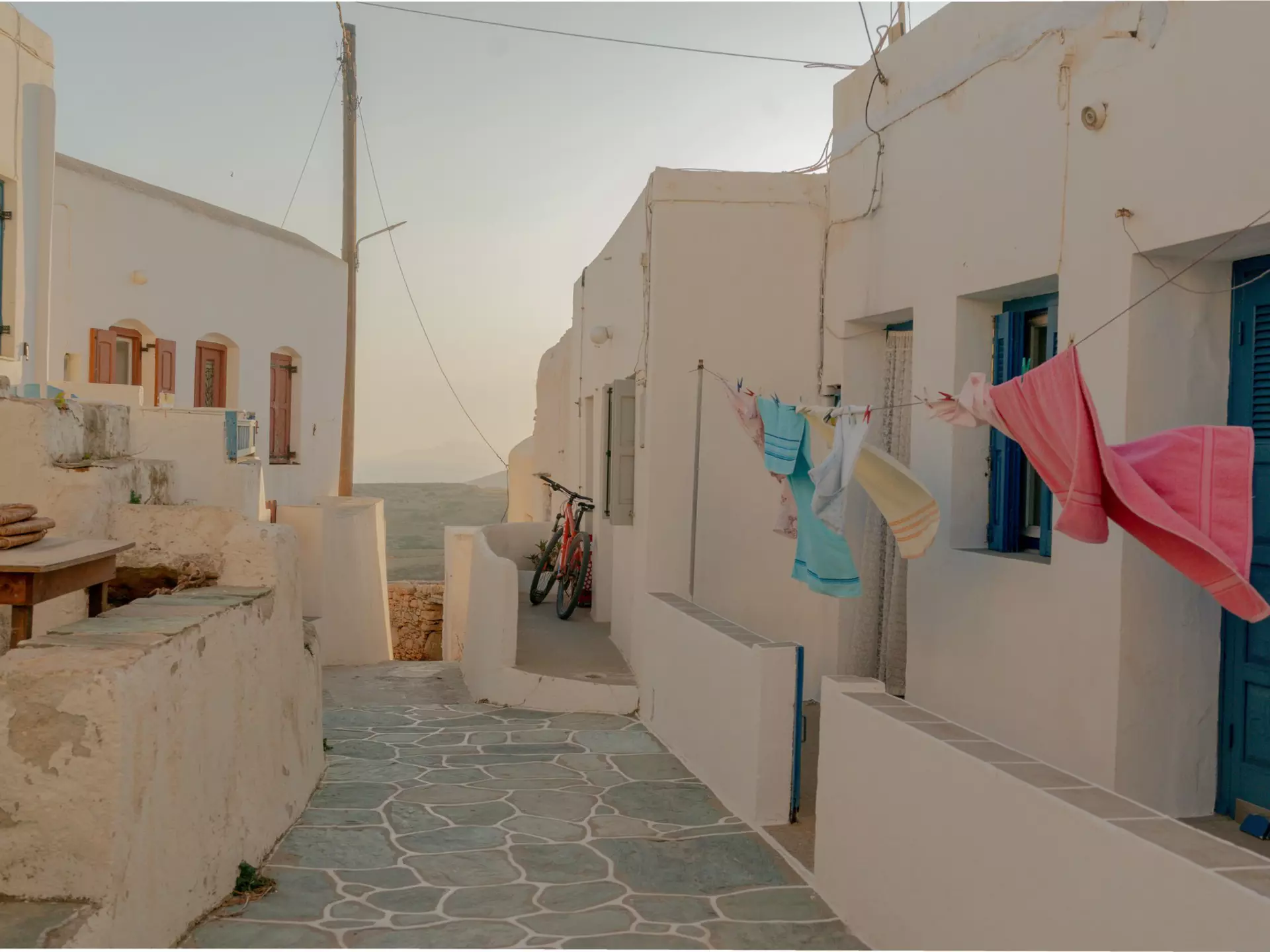 Colorful towels hang on the line outside a white building on a narrow cobblestone walkway in the Cyclades, Greece; the sea is visible at the end of the alley.