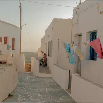 Colorful towels hang on the line outside a white building on a narrow cobblestone walkway in the Cyclades, Greece; the sea is visible at the end of the alley.