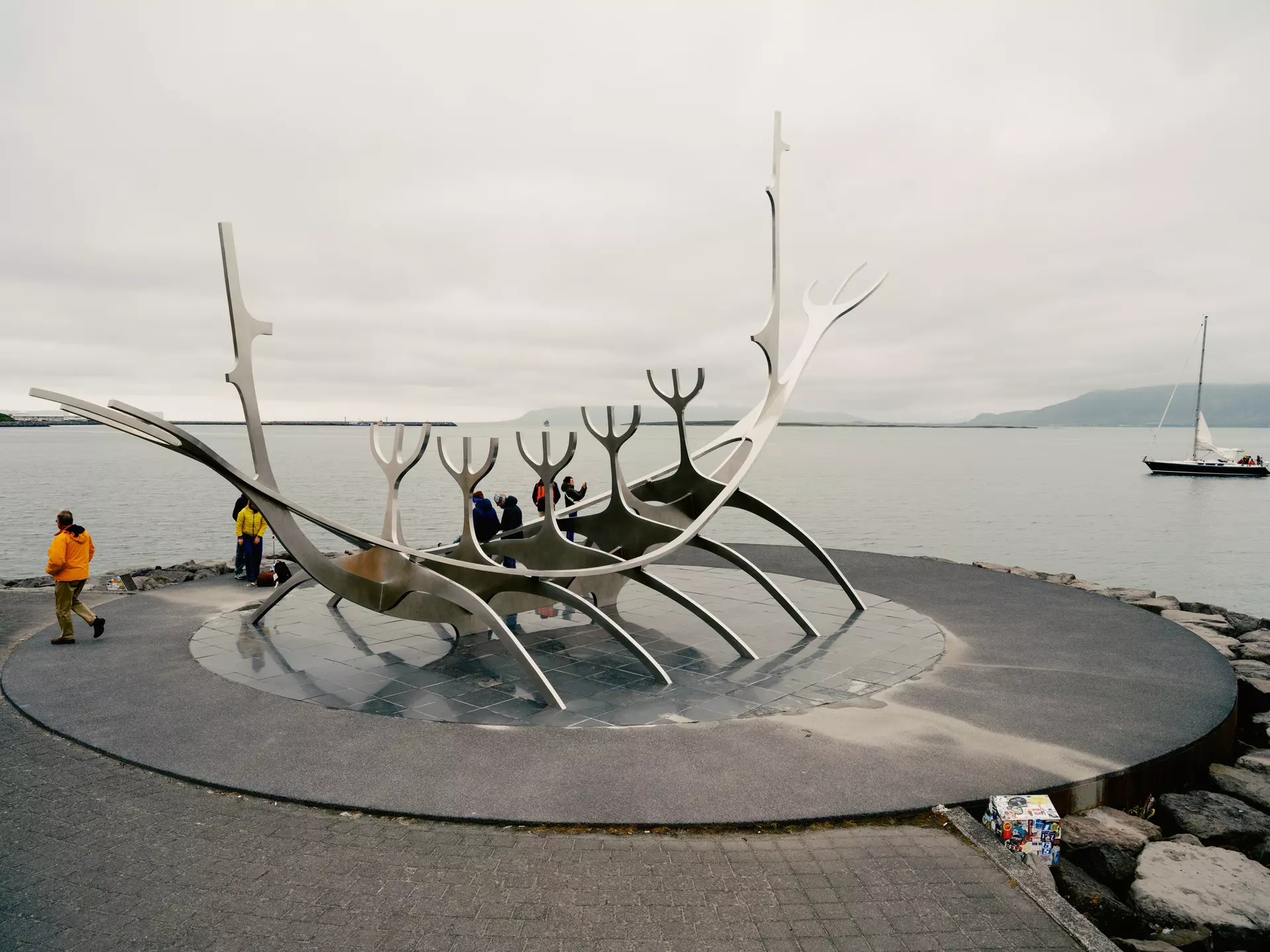 A large, steel public sculpture that resembles a long ship on a shore. People stand nearby taking photos.