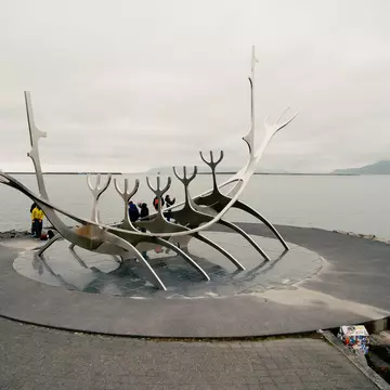 A large, steel public sculpture that resembles a long ship on a shore. People stand nearby taking photos.