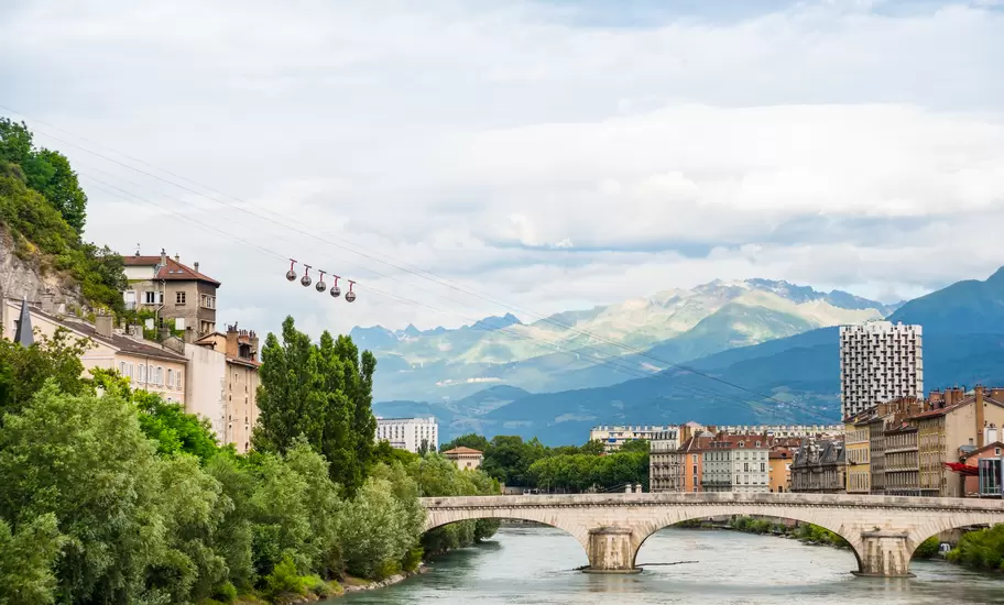 Grenoble's Bastille cable-car bubbles above the Isère River and a stone bridge, with Alpine peaks in the distance