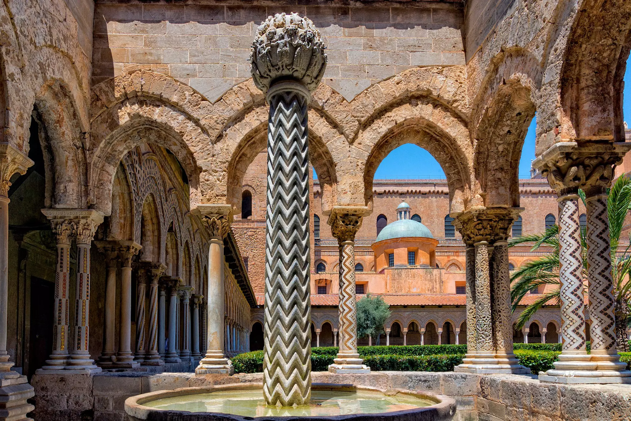 A courtyard with mosaic-covered columns and stone archways with a reddish stone building in the distance on a sunny day.